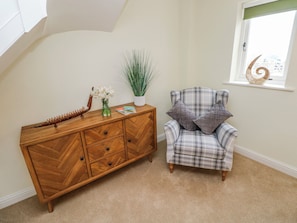 Une pièce comprenant une commode en bois à motifs en chevrons, un fauteuil à carreaux, une fenêtre ornée d’une coquille décorative et une plante.