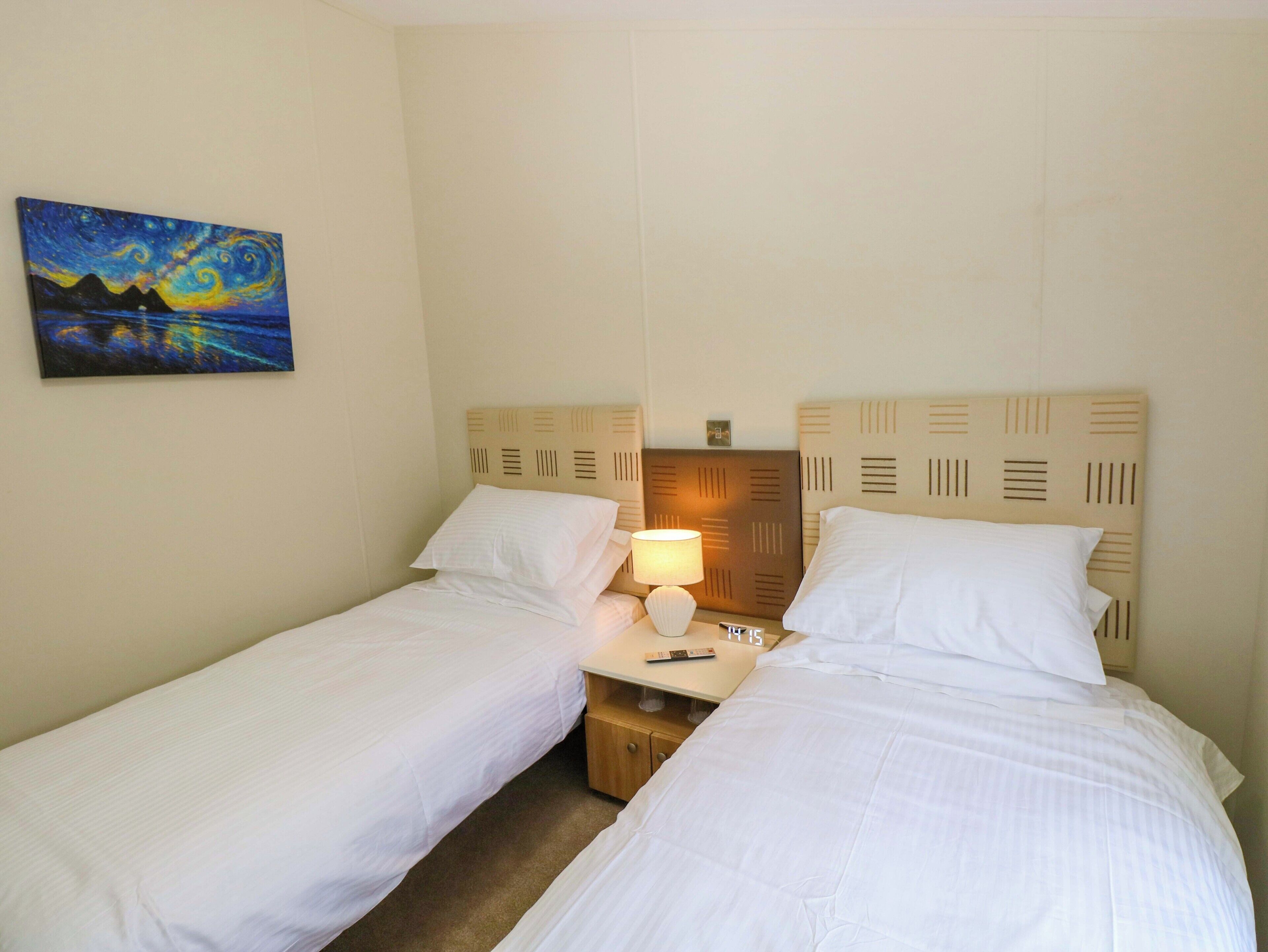 A hotel room with two single beds, a nightstand with a lamp, a painting on the wall, and a clock.