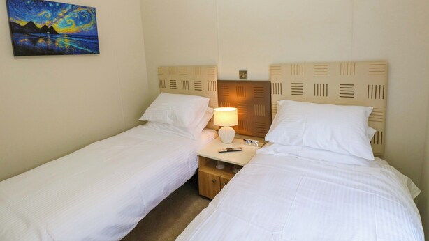 A hotel room with two single beds, a nightstand with a lamp, a painting on the wall, and a clock. - Wernlas Lodge (Swansea)