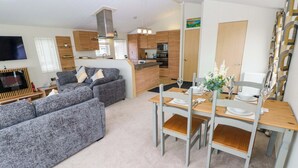 A modern living room with a grey sofa set, a dining table set for four, and a kitchen area with wooden cabinets and a microwave. - Wernlas Lodge (Swansea)