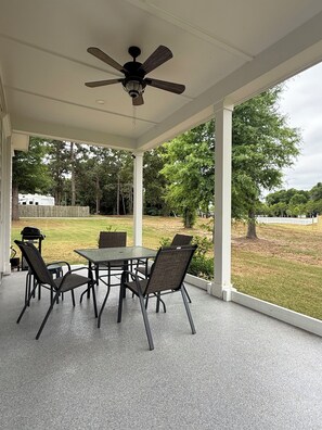 A covered patio with a table and chairs, a ceiling fan, and a view of a grassy yard with trees.