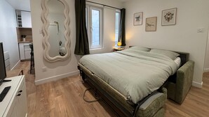 A bedroom with a bed, a mirror, a window with curtains, a bedside table with a lamp, and a small kitchenette. - Modern, Renovated Studio in the Heart of La Rochelle by Kaz'Concept (La Rochelle)