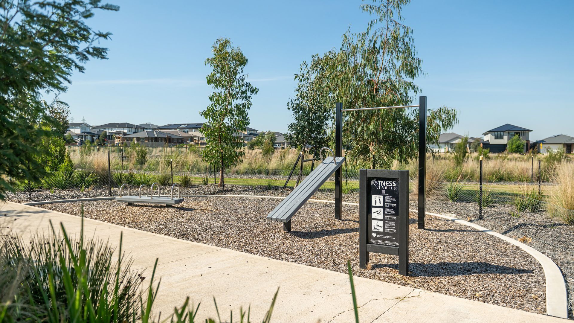 A fitness trail sign, exercise equipment, and a playground area in a park setting.
