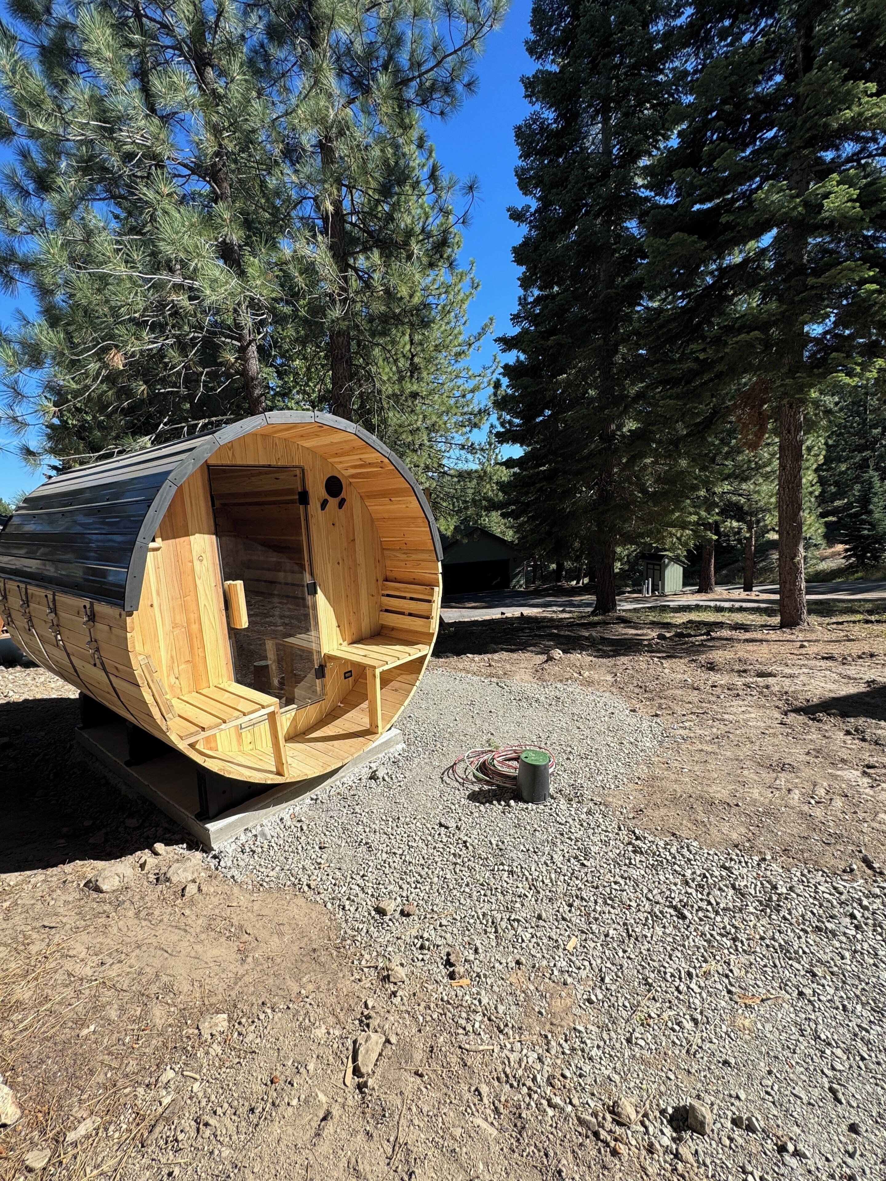 A wooden cylindrical structure with a bench inside, set on a gravel surface with trees in the background.