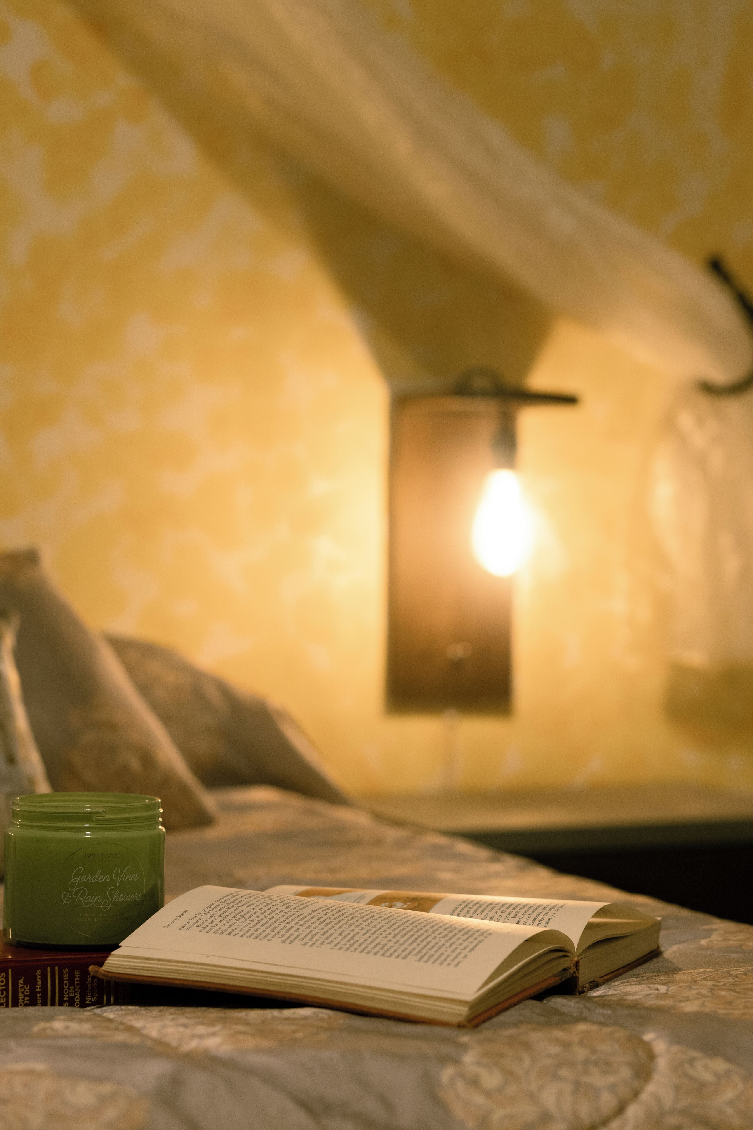 Un lit avec un livre et une bougie, une lampe fixée au mur et un mur décoré d’un motif.