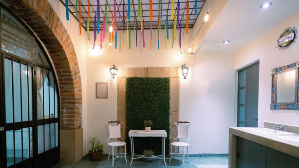Una habitación con pared de ladrillo, pared verde, mesa blanca con dos sillas, un lavabo y cintas de colores colgando.