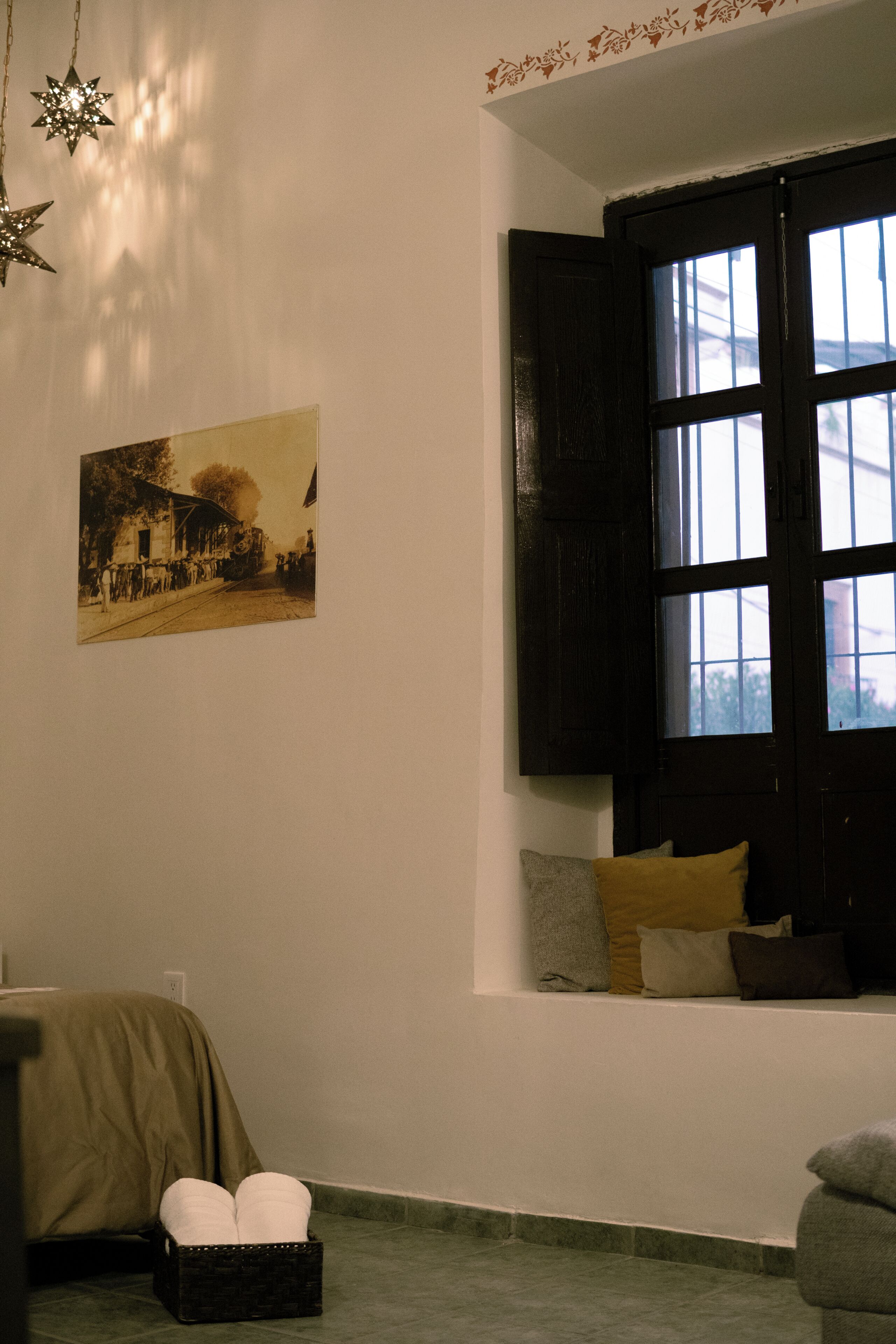 A room with a large window, a wall-mounted picture, a bed with a blanket, and a bedside table with a lamp.