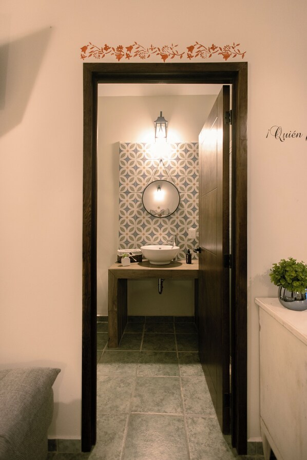 Une salle de bain avec une porte en bois, un lavabo avec un miroir, un papier peint à motifs et une petite plante posée sur le comptoir.