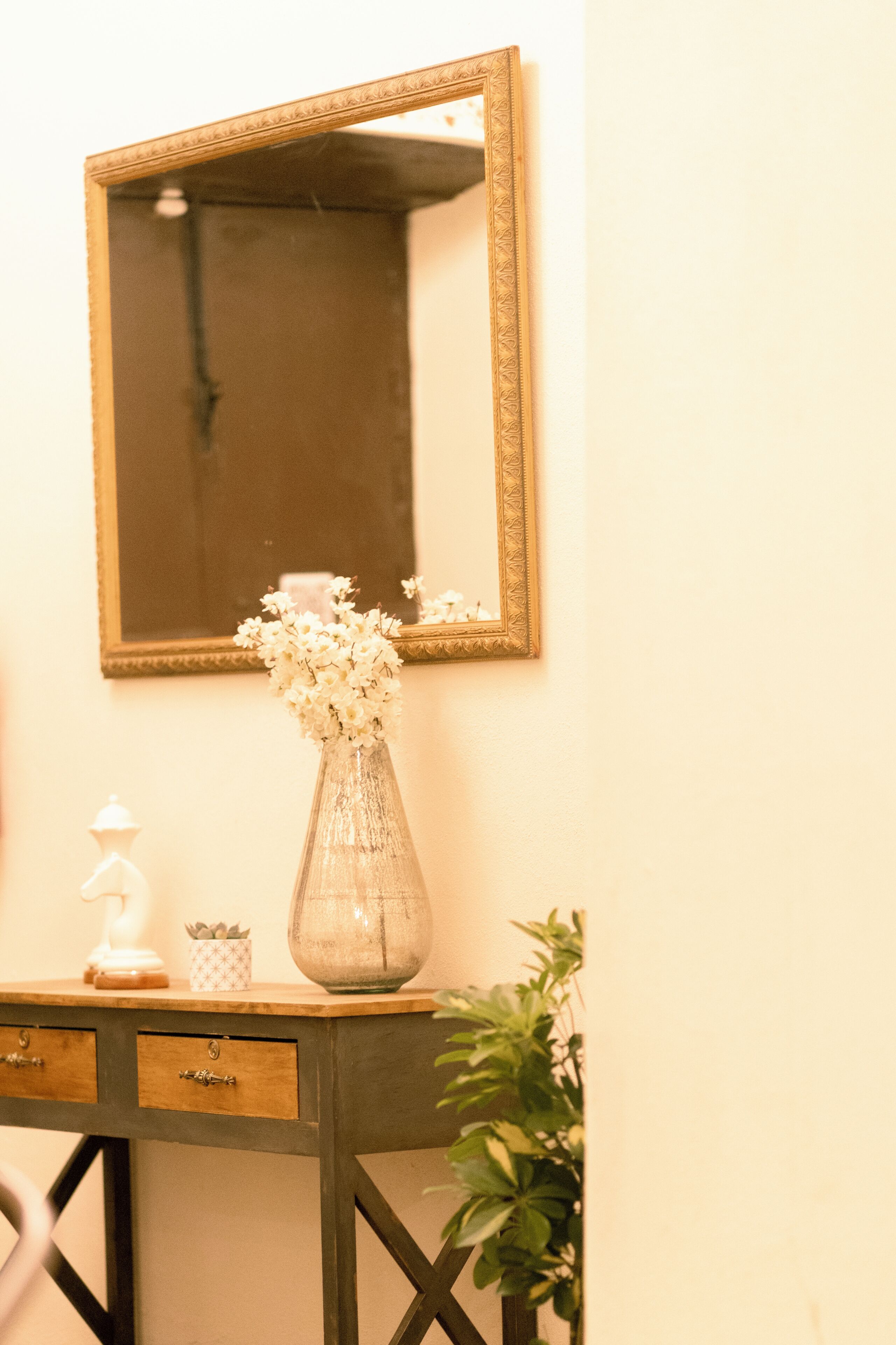 A mirror on a wall, a vase with flowers, a small plant, and a wooden console with drawers.