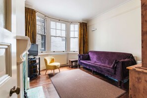 Ein Zimmer mit einem violetten Sofa, einem gelben Sessel, einem kleinen Tisch und einem Fernseher auf einem Schrank.