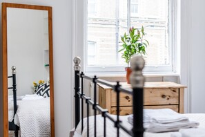 Ein Schlafzimmer mit einem Bett, einer Holzkommode, einem Spiegel und einem Fenster mit Blick auf Gebäude.