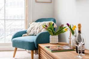 Ein blauer Sessel mit weißer Decke, ein Holztisch mit einem Buch, eine Vase mit Blumen und eine Weinflasche.