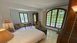 Ein Schlafzimmer mit einem großen Bett, einem Schreibtisch, einem Stuhl und zwei Fenstern mit Blick auf Grünflächen.