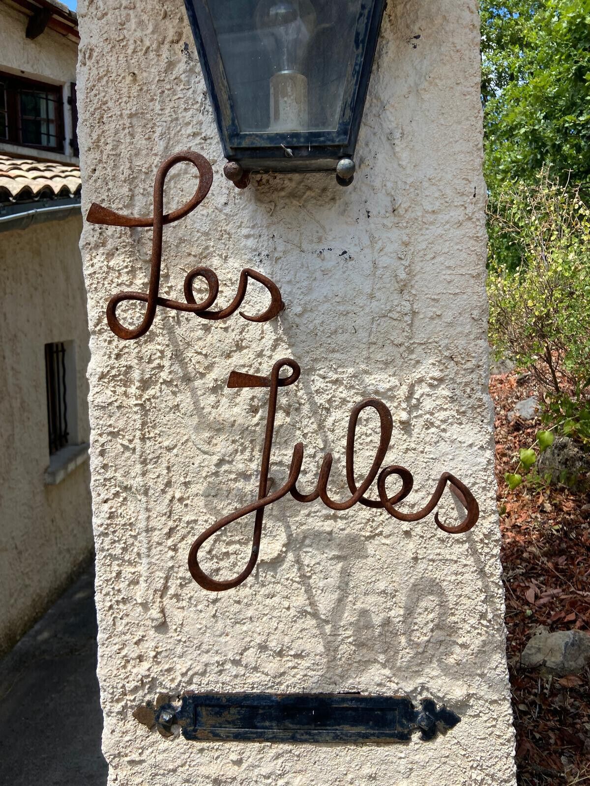 A wall with the inscription 'Les Jules' in a cursive script, a black metal lantern above it, and a small metal plaque below.