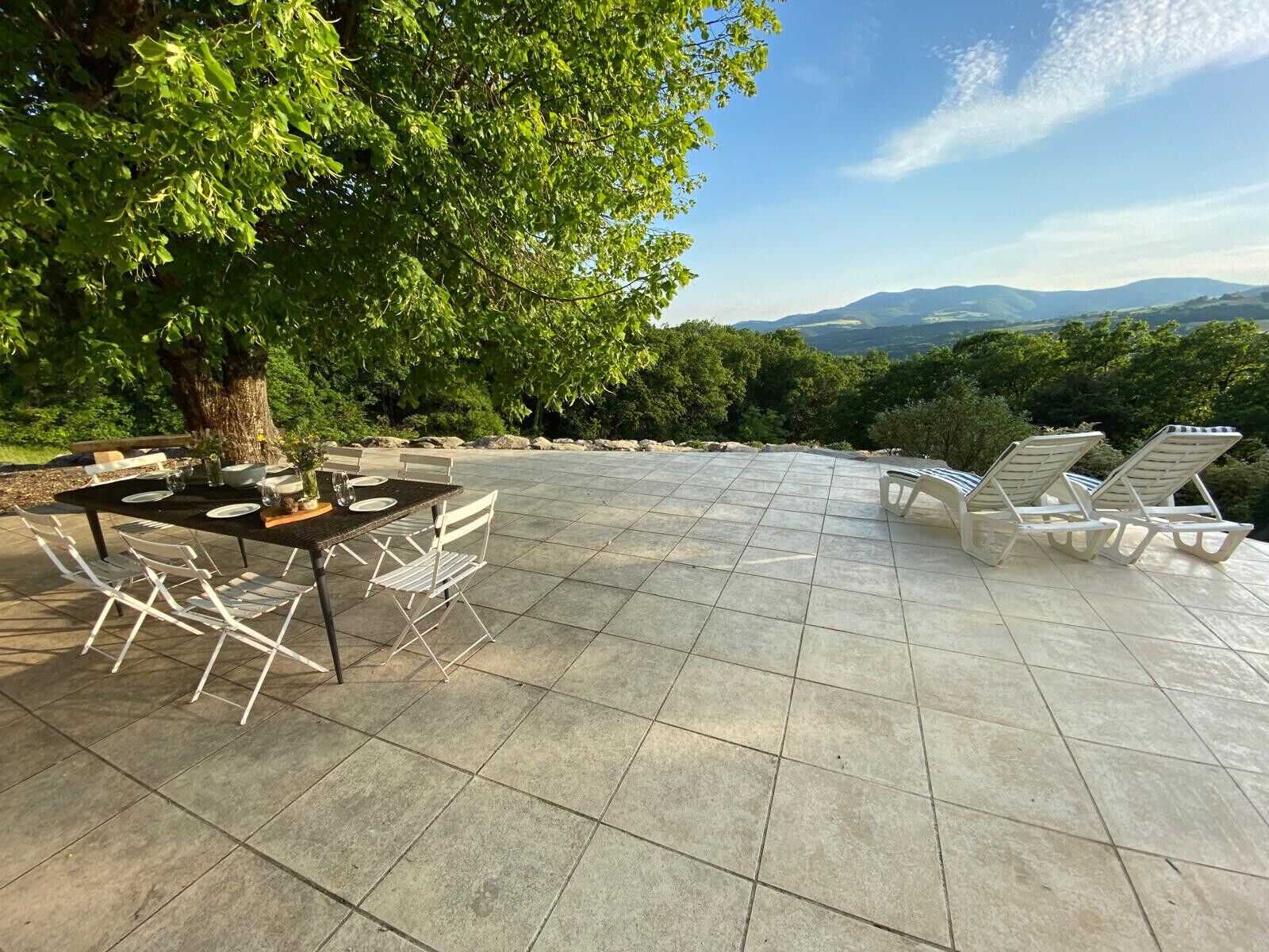 Une terrasse extérieure avec une table dressée pour les repas, entourée de fauteuils confortables et un grand arbre qui offre de l’ombre.
