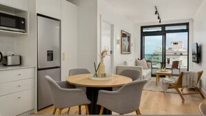 A modern kitchen with white cabinetry, a built-in microwave, and a refrigerator. There is a dining area with a round table and chairs, and a living space with a sofa and a TV. - Sage Sea Point 1 Bed Ocean View (Cape Town)