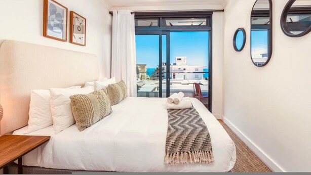 A hotel room with a bed, a wooden nightstand, a large window with a view of buildings, and two framed pictures on the wall. - Sage Sea Point 1 Bed Ocean View (Cape Town)