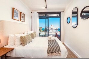A hotel room with a bed, a wooden nightstand, a large window with a view of buildings, and two framed pictures on the wall. - The Sage Apartment 607 (Cape Town)
