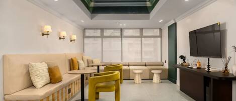 A modern hotel lobby with a sofa, yellow chairs, a small table, a TV, and a reception desk.