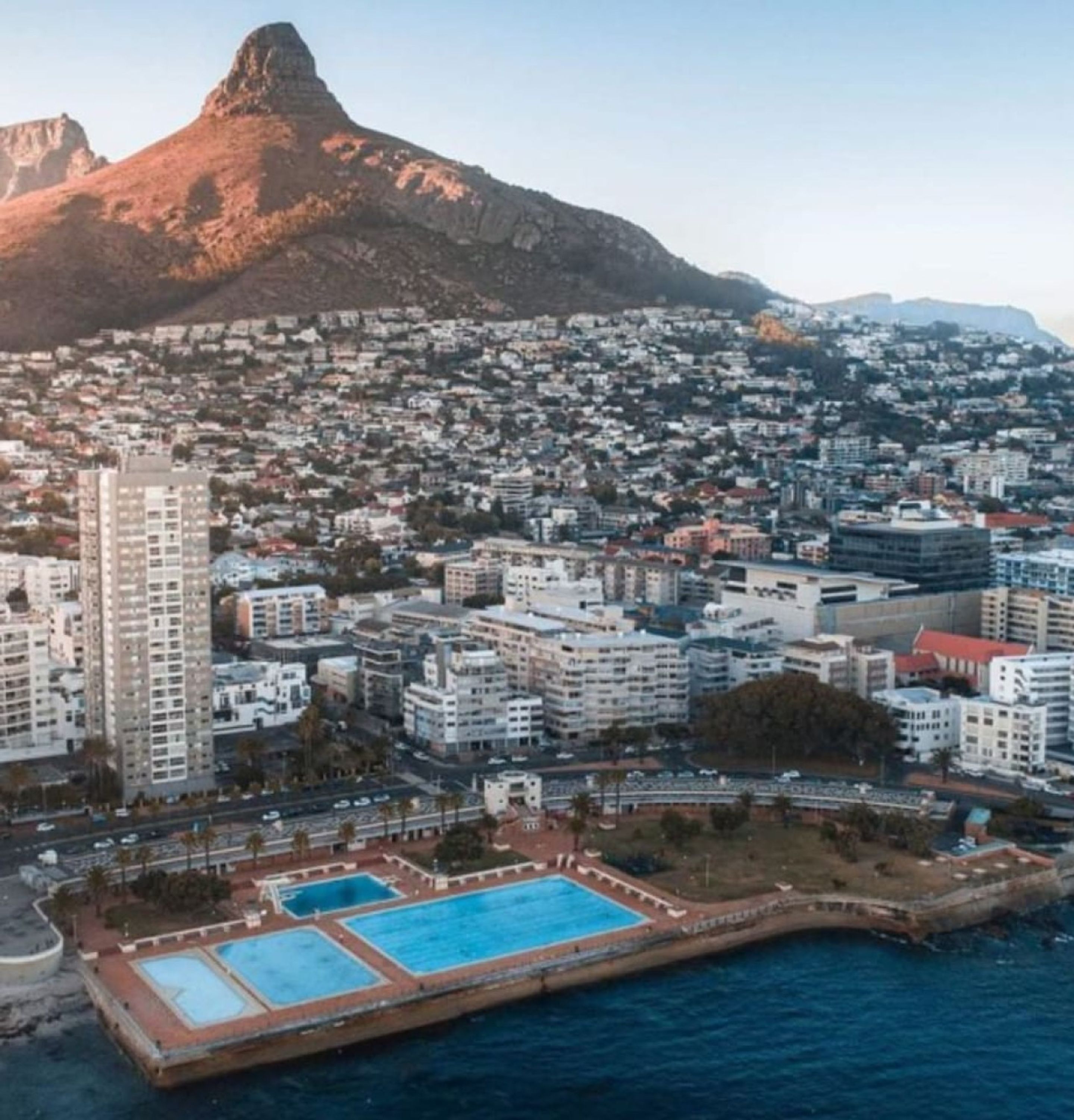 A coastal cityscape with a prominent mountain in the background, a large hotel with multiple floors, and a beachfront swimming area.