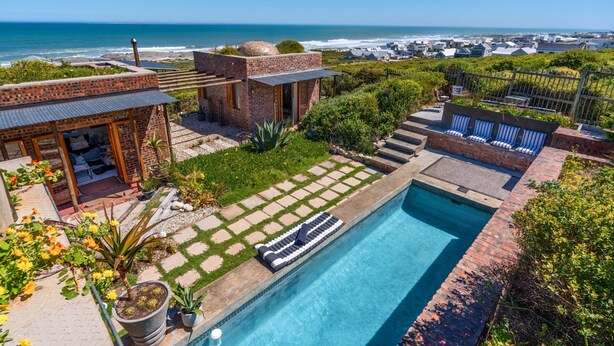 A pool with a view of the ocean, a patio area with lounge chairs, and a brick building. - Yzerfontein 3 Bed House With Pool Sea Views (Yzerfontein)