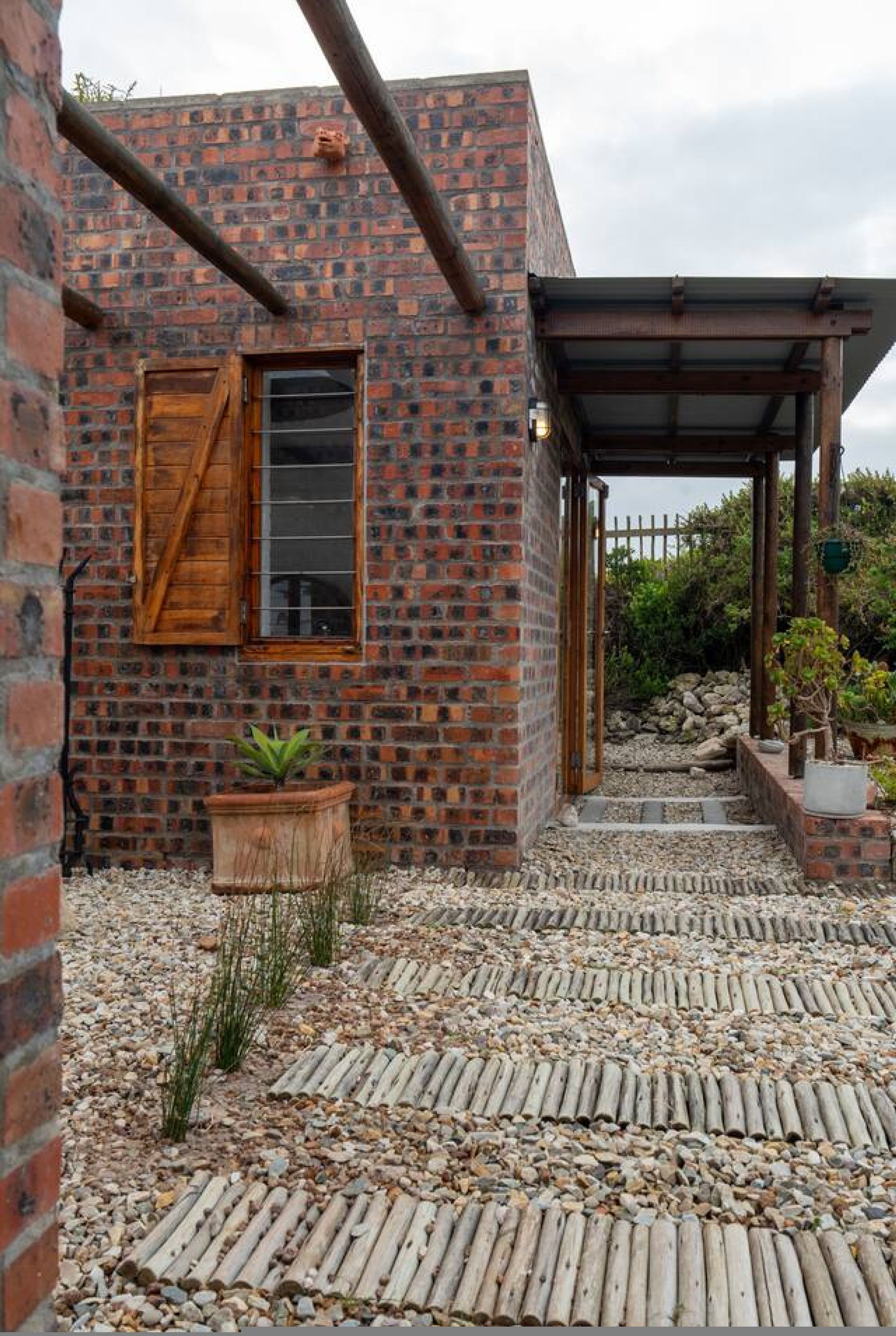 A brick building with a wooden door, a potted plant, and a gravel pathway leading to a covered area.