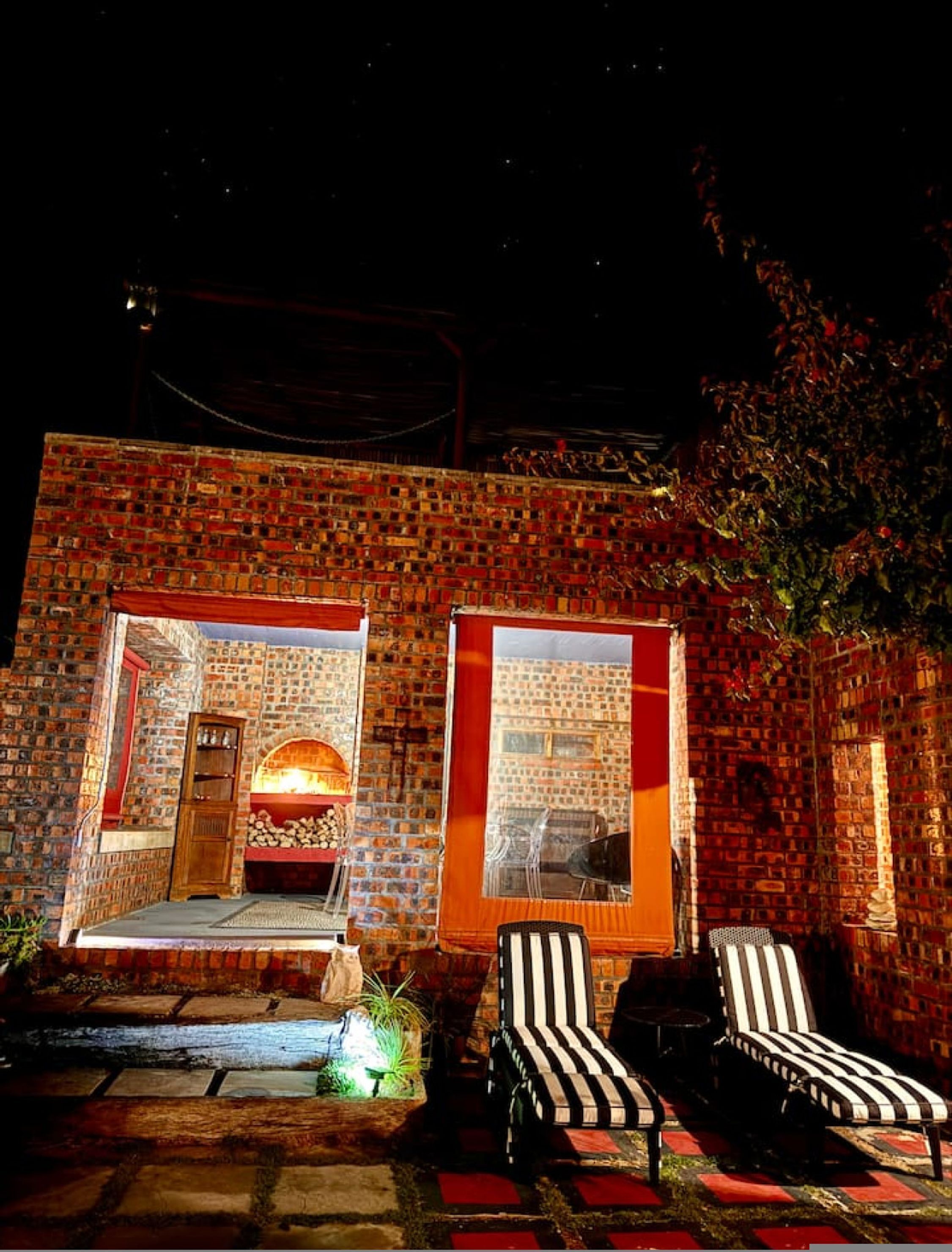 A brick building with two large windows, a fireplace, and two striped lounge chairs outside.