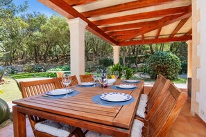 Ein Holztisch im Freien, gedeckt für ein Essen, mit Stühlen, umgeben von einem Garten mit Bäumen und einer Steinmauer.