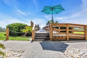 A paved pathway leading to a wooden bridge with a blue umbrella, surrounded by greenery and a clear sky. - Ocean Front Condo- Indoor & Outdoor Pools- Lazy River + Hot Tub (North Myrtle Beach)