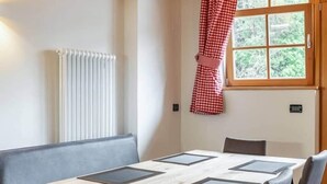 Une pièce avec un plafond en bois, un radiateur, une fenêtre ornée de rideaux à carreaux rouges, une table dont le plateau est de couleur claire et sur laquelle reposent des sets de table carrés de couleur foncée, et des sièges recouverts d’un tissu gris.