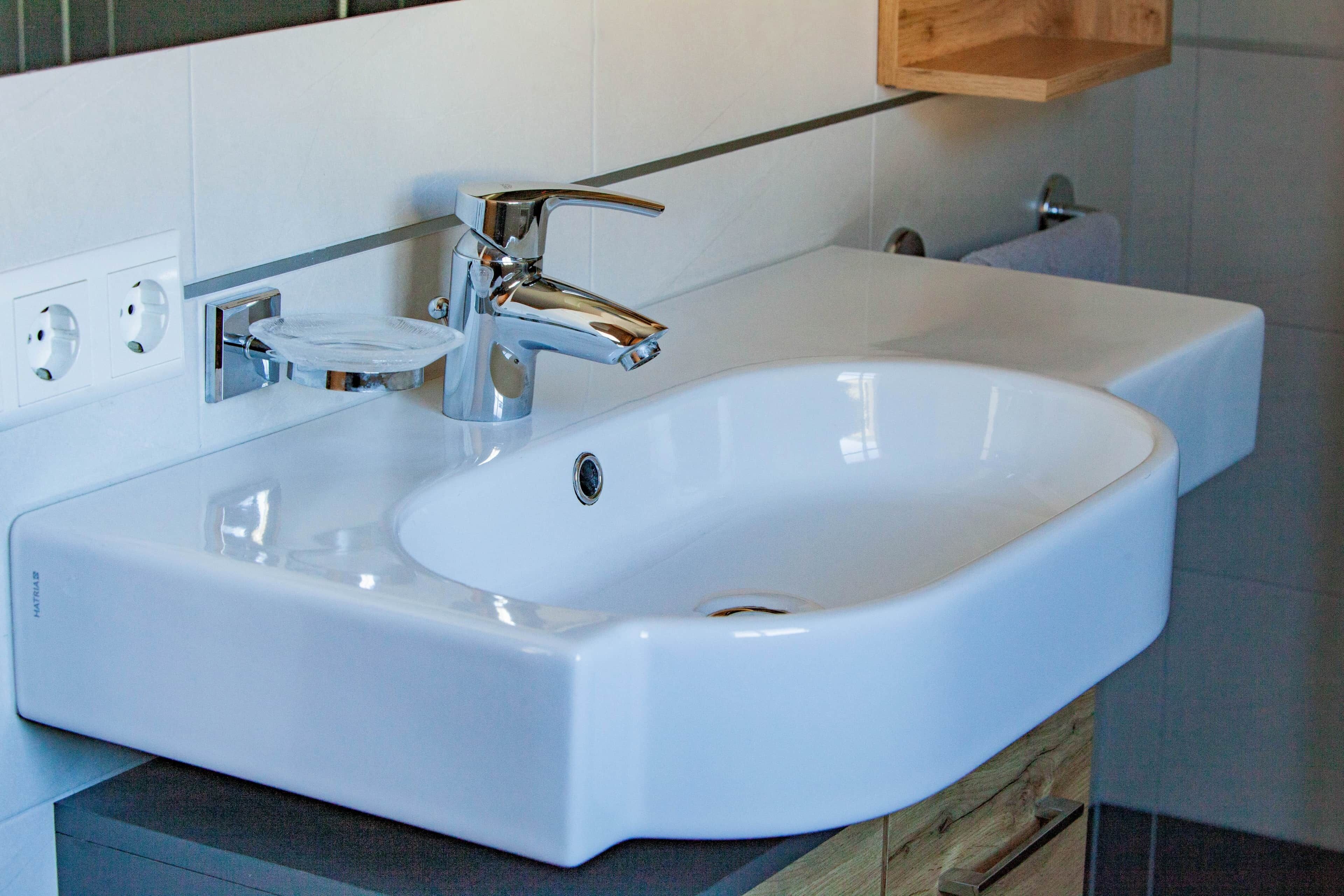 Ein weißes Waschbecken mit chromfarbenen Armaturen, eine Seifenschale und ein Holzregal darüber.