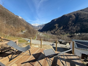 Une terrasse en bois avec deux chaises longues, une vue sur une vallée bordée de montagnes et une route qui s’éloigne à perte de vue.