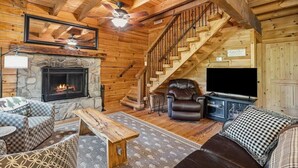 L’intérieur d’une cabane en rondins, avec une cheminée en pierre, un escalier en bois, des sièges en cuir et un téléviseur.