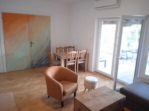 A room with a wooden dining table, chairs, a sofa, a small round stool, and a sliding glass door leading to an outdoor area.