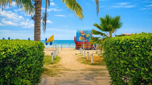 Uma trilha na praia que leva a um mural colorido, uma bandeira e um restaurante à beira-mar.