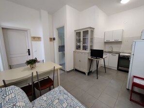 Eine kompakte Küche mit weißen Schränken, einem Esstisch mit Stühlen, einem kleinen Fernseher auf einem Standfuß und einem Kühlschrank.