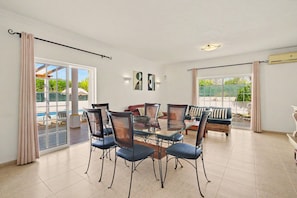 A spacious room with a glass dining table surrounded by chairs, large windows with curtains, and a view of the outdoors.