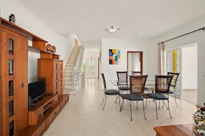 A spacious living area with a wooden entertainment unit, a dining table with chairs, a staircase, and a large mirror.