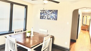 A dining area with a wooden table and white chairs, a ceiling fan, a window with blinds, and a painting on the wall.