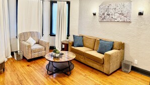 A living room with a patterned armchair, a sofa with blue cushions, a round coffee table, and a painting on the wall.