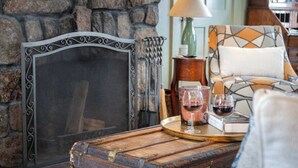 Une pièce chaleureuse avec un mur en pierre, un paravent à ossature métallique, une table en bois sur laquelle repose un plateau, des verres de vin rouge et un fauteuil à motifs.