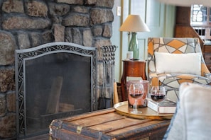 Een gezellige kamer met een stenen muur, een scherm met metalen frame, een houten tafel met dienblad, glaasjes rode wijn en een gestreept fauteuil.