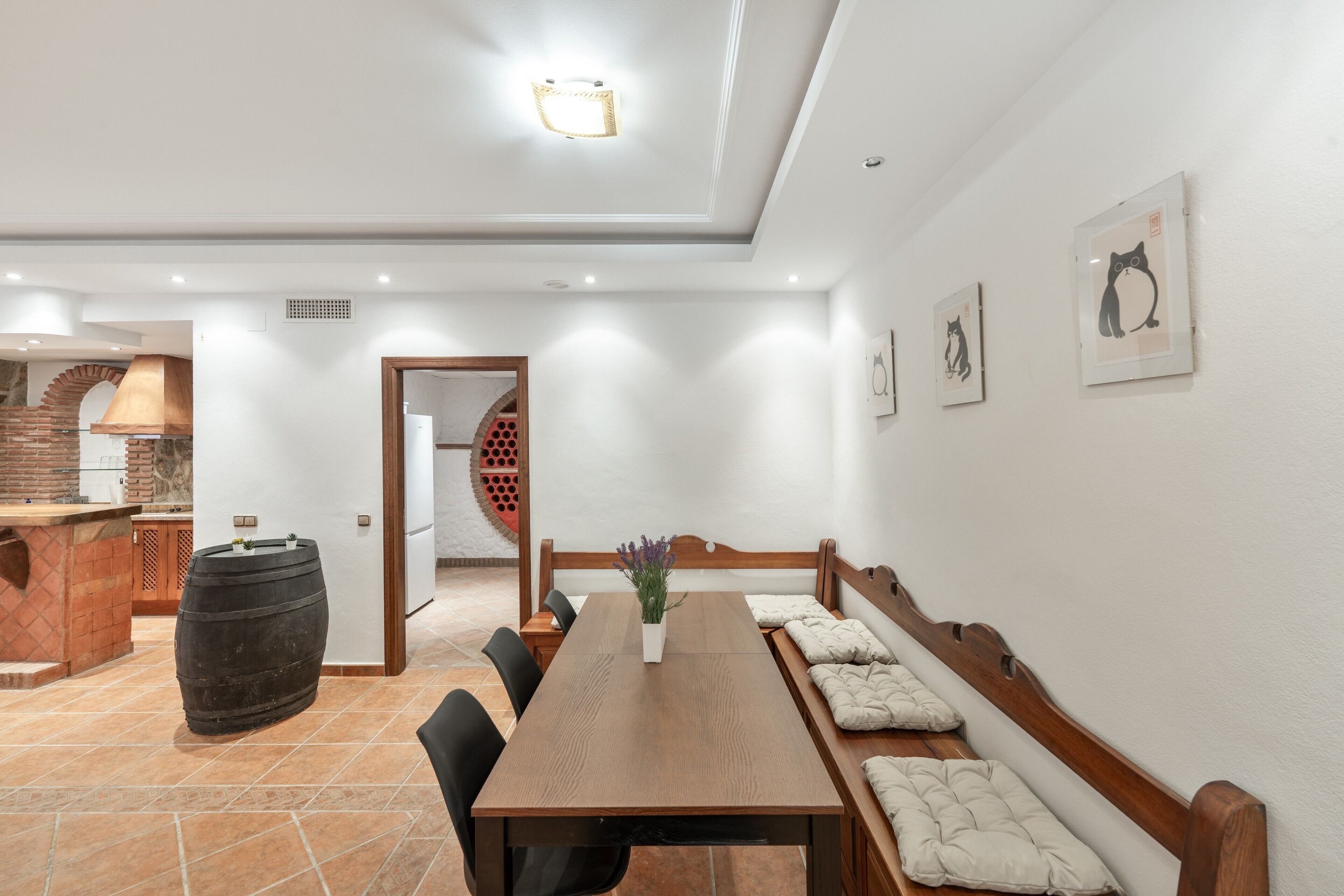 Un comedor con mesa de madera, sillas y un banco con cojines. Hay un barril, una planta en maceta y obras de arte enmarcadas en las paredes.