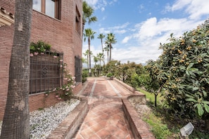 A brick building with a tiled pathway, palm trees, and a bush with yellow fruits.