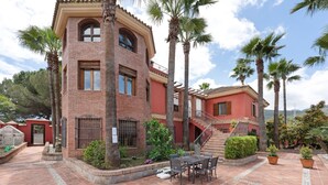 Ein zweistöckiges Gebäude mit roter Fassade, einem Backsteinbereich und einem Balkon. Eine Terrasse mit Tisch und Stühlen ist von Palmen und Topfpflanzen umgeben.