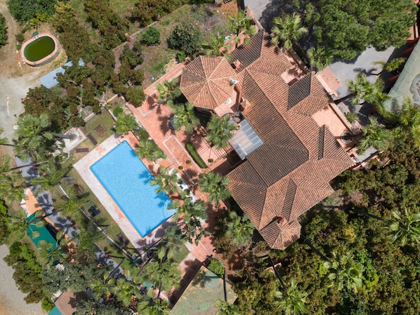 Vue aérienne d’une propriété dotée d’une piscine, d’un toit recouvert de tuiles et entourée de verdure.