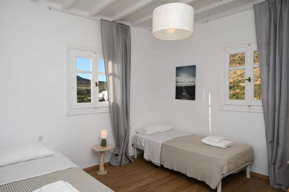 Un dormitorio con dos camas, una mesita de noche con lámpara, una ventana con cortinas y un cuadro en la pared.