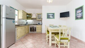 A compact kitchen with a refrigerator, microwave, oven, and wooden cabinets. There is a dining table with chairs and a flat-screen TV mounted on the wall. - Sardinian Apartment for 6 with Private Terrace, Three Minutes’ Walk from the Beach (Orosei)