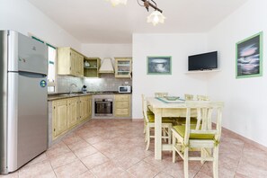 Cocina compacta con refrigerador, microondas, horno y gabinetes de madera. Hay una mesa de comedor con sillas y un televisor de pantalla plana montado en la pared.