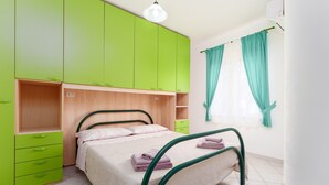 A room with a bed, green cabinets, a wooden shelf, and a window with curtains. - Sardinian Apartment with Private Terrace 3 Minutes’ Walk from the Beach (Orosei)
